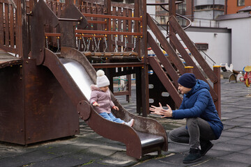 Father catching his toddler daughter sliding down a playground slide