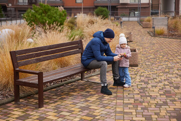 Father and daughter interacting with mobile phones while sitting on a bench in an urban park