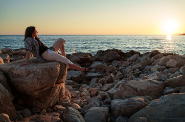 A girl poses and relaxes by the sea, sitting on rocks near the slope of a high cliff, panoramic view of the seashore and mountains at sunset