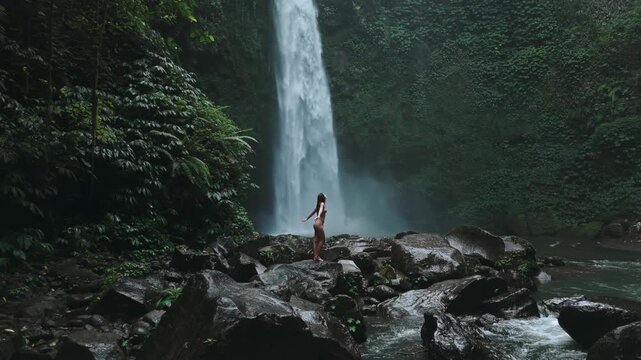 A mesmerizing view of a tall waterfall cascading into a lush tropical ravine in Sekumpul, Bali. An observer stands near the stream, embracing nature's grandeur.