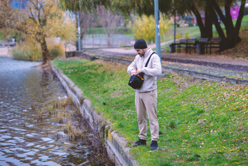Fototapeta premium A man in a beanie and fleece jacket stands by a lake on a cool autumn day, looking into his shoulder bag. Serene outdoor scene capturing a moment of contemplation or preparation in nature.