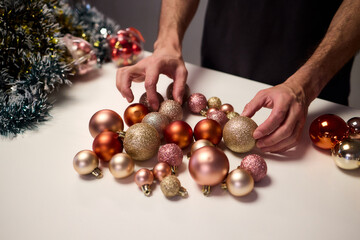 Hands arranging Christmas New Year baubles on white table with tinsel and warm bokeh lights, metallic red gold pink palette, careful styling by home decorator, festive craft mood