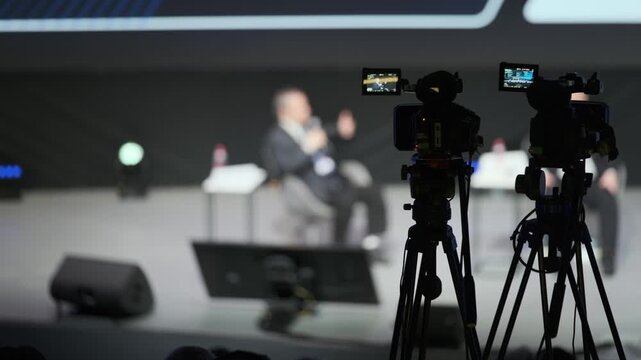 Blurred stage interview session with cameras in foreground and audience unseen.