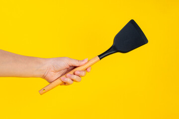 Hand with Black plastic kitchen spatula, kitchenware for cooking on yellow background.