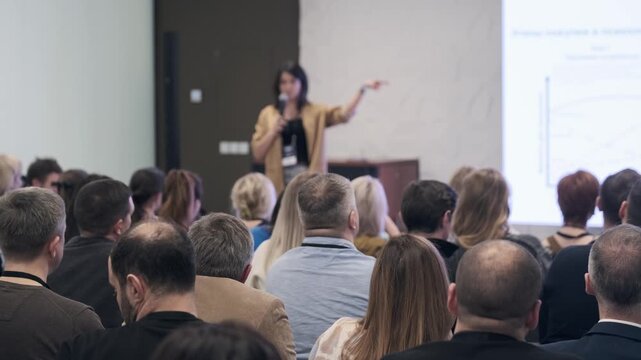 Presenter speaks to crowded room during training session audience listens notes focus leadership communication industry insights in dynamic conference setting.