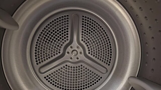 Inside view of a clothes dryer drum with metal surface and ventilation holes. Household laundry appliance interior shown in clean technical detail.