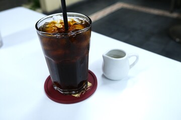 Refreshing iced black oolong tea in a tall glass with a straw, accompanied by a small creamer pitcher on a clean white table.
