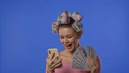 A woman with curlers wrapped in money holds a fan of banknotes, looks at her smartphone screen, and...