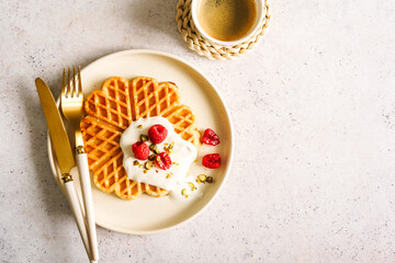 Gebackene Waffeln mit Joghurt und frischen Himbeeren auf einem beigen Teller und eine Tasse Kaffee auf einem grauen Tisch. Draufsicht, Fr&uuml;hst&uuml;ck.