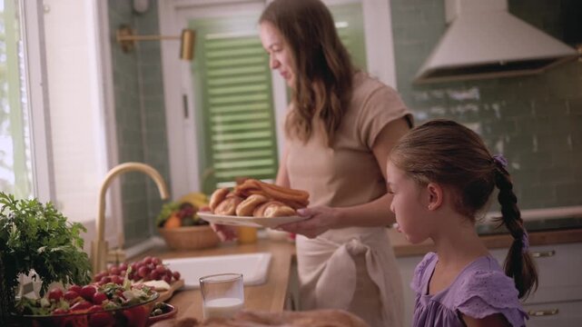Joyful little girl playfully dipping a traditional spanish churro into a glass of milk while her smiling mother watches, sharing a happy family moment together during breakfast at home