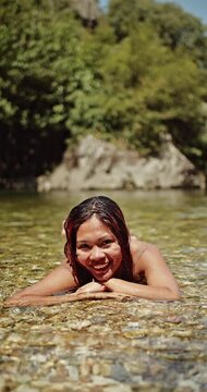 A cheerful woman relaxes in the crystal-clear water of a natural river, her wet hair glistening as she smiles brightly. Perfect for showcasing summer relaxation and outdoor activities.