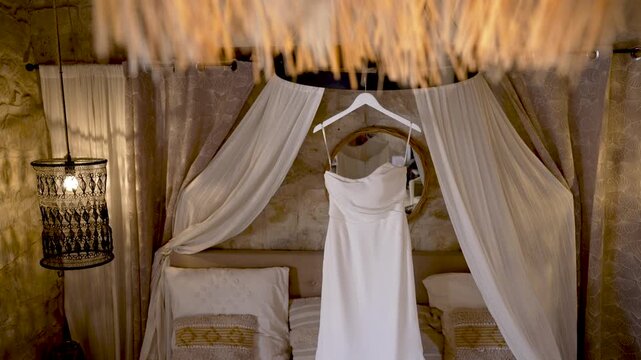 A stunning white wedding dress hangs gracefully in an inviting room, surrounded by warm lighting and elegant furnishings, perfect for a memorable occasion.