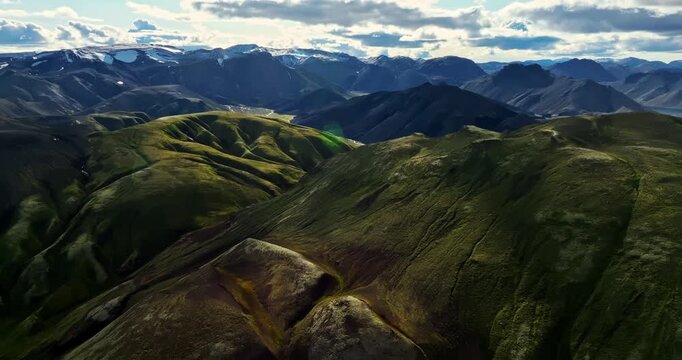 Rolling green ridges unfold toward distant icy peaks, capturing Iceland&rsquo;s rugged highlands with soft sunlit contours and deep shadowed valleys from a high-elevation aerial perspective.