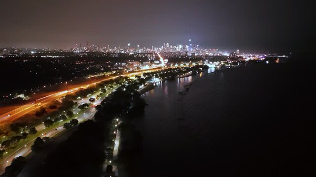 Nighttime aerial footage overlooking Lake Ontario with the Gardiner Expressway and Lakeshore Boulevard glowing toward the illuminated downtown Toronto skyline.