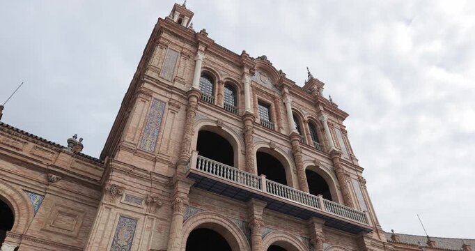 Low-angle shot of a tower section of the Plaza de Espa&ntilde;a building in Seville, Spain, featuring Neo-Moorish architecture, ornate arches, columns, and ceramic tilework under a cloudy sky