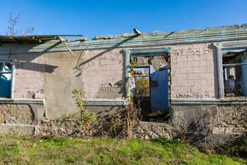 Old, dilapidated building with broken roof and walls, blue sky in the background, overgrown grass and plants in front, symbolizing decay and abandonment.