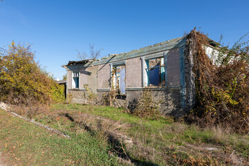 Old deserted house with peeling paint and overgrown vegetation under a clear blue sky, evoking themes of neglect and decay.