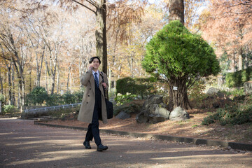 秋の紅葉の屋外でコートを着て営業の電話など通話をする男性	全身広角