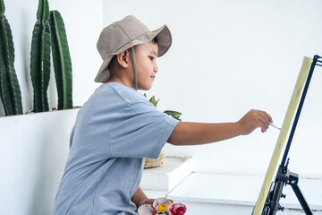 Side View Of Young Asian Boy Artist Wearing Bucket Hat Painting On Canvas