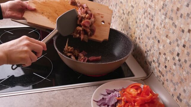 Home cook slides marinated meat pieces from a wooden cutting board into a preheated wok on the stove, first step of cooking a hot stir fry or stew, ideal for recipe videos