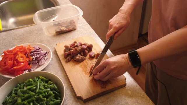 Woman slicing marinated meat on a wooden cutting board next to bowls of chopped red pepper, red onion, and green beans, preparing ingredients for a hearty hot dish