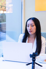 Asian woman working remotely in cafe using laptop and tripod