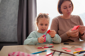 Mother and her little daughter cut out paper hearts sitting at a desk. Family is preparing for the celebration of Valentine's Day