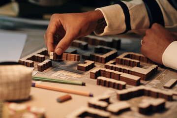 Fototapeta premium Young Black man arranging architectural model pieces on table, hand placing miniature building on urban planning layout, visible tattoo on wrist, pencils and eraser nearby