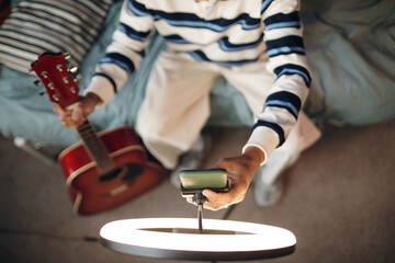 Young Black man sitting on bed holding acoustic guitar in one hand and adjusting ring light with other hand, preparing for music recording or online streaming session at home