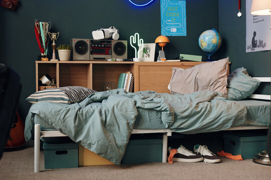 Messy bed with striped pillow and wrinkled blanket in teenager bedroom, bookshelf holding trophies, books, virtual reality headset, radio, globe, sneakers and boxes under bed