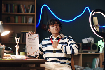 Black teenager sitting at desk holding certificate of achievement and showing gold medal around neck, bookshelf and trophy in background, looking confidently into camera during social media livestream