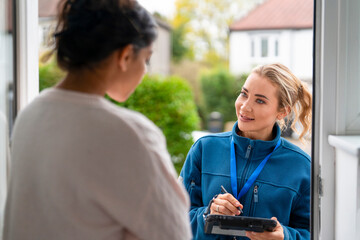 Woman talks with another woman at the door while holding a tablet in a residential area during...