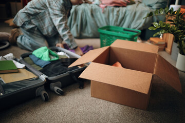 Young woman kneeling on floor packing suitcase with clothing and personal items, open cardboard box...