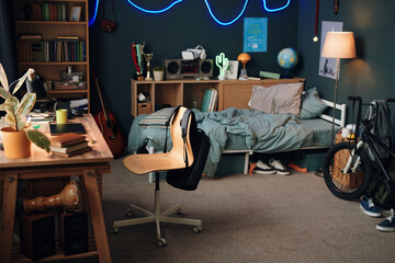 Teenager bedroom interior showing unmade bed, wooden desk with books and plant, bicycle leaning against wall, various personal items and decorations scattered throughout space © DragonImages