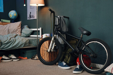 Teenager bedroom showing stunt bicycle leaning against wall near unmade bed, sneakers on floor, backpack and globe in background, suggesting active lifestyle and youthful environment