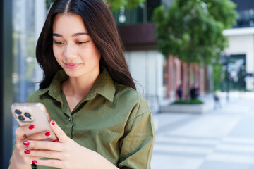 Young, beautiful Asian woman in green shirt smile, using smartphone in front of modern office building. Digital technology, online shopping lifestyle, social media and communication concept
