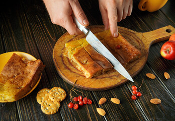 A person cuts a homemade dessert on a wooden board. Nearby are slices of apples, nuts, and crackers. The scene captures a warm, inviting kitchen atmosphere