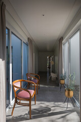  Bright, modern residential corridor flooded with natural light, featuring elegant wooden chairs with pink upholstery, contemporary gold planters, and large windows revealing an outdoor view