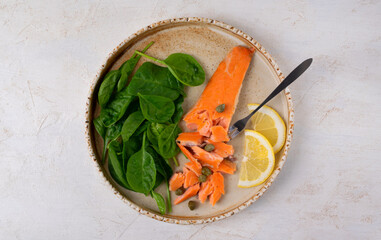 Hot smoked trout fillet, finely crumbled with a fork, served with spinach, lemon and capers on a ceramic plate. Top view. Copy space on a white background