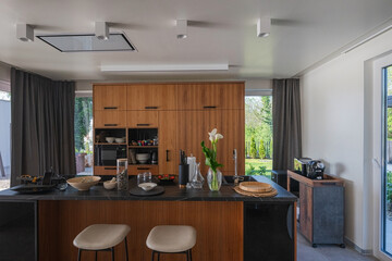 Modern luxury kitchen interior with wooden cabinetry, central island with bar stools, and ambient LED lighting. Stylish contemporary design with large windows and dark countertops at twilight