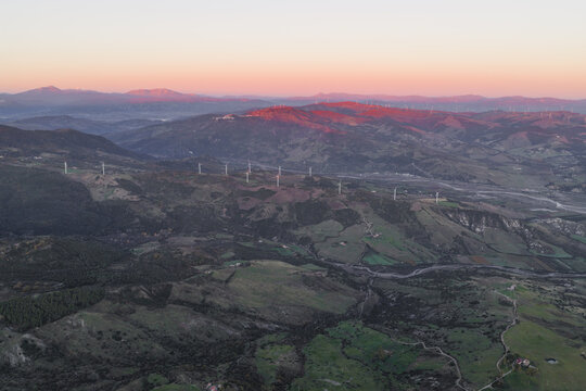 Aerial view of the sun kissing the wind turbines atop rolling green hills, a symphony of nature and technology harmonizing under a pastel sky, Rotondella, Basilicata, Italy.