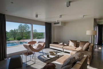 Obraz premium Modern luxury living room interior at sunset. Open-plan space featuring beige sofas, marble dining table, large glass windows with sun flare, and view of a patio. Stylish contemporary home design