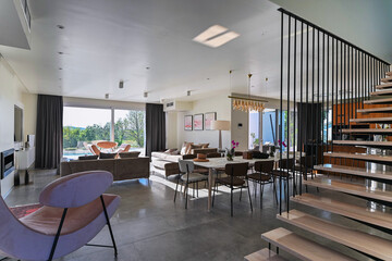 Modern luxury open-plan interior featuring a floating staircase, marble dining table, wooden kitchen, and beige sofa. Spacious contemporary home design with elegant lighting and gray tiled floors