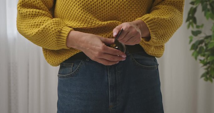 A woman fastens a leather belt on her jeans, wearing a yellow knitted sweater, close up of her waist indoors.