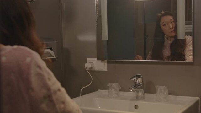 An attractive Asian woman brushes her hair while looking in the mirror of a hotel bathroom