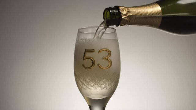 Glass engraved with the number 53. Champagne bottle fills the glass with alcohol. Celebrating milestone, anniversary or birthday