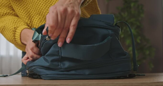 Woman in a yellow sweater unzipping a dark blue backpack on a table, close up of hands and travel gear