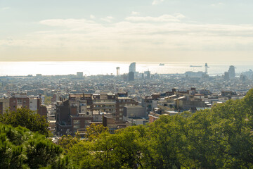 View of sprawling cityscape meets the shimmering sea under a soft sky, green trees framing the urban vista, contrasting nature with the architectural skyline, Barcelona, Catalonia, Spain.