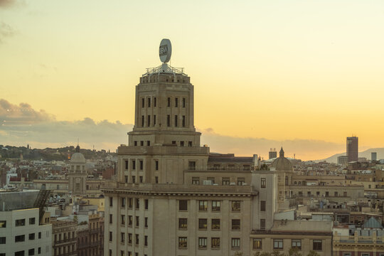 View of a stately building crowned with a distinguished emblem, standing tall amidst the urban expanse under a warm sunset glow, Barcelona, Catalonia, Spain.