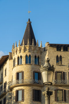View of a building's tan stone facade with sharp dark roofs and a golden light fixture under a clear blue sky, Barcelona, Catalonia, Spain.